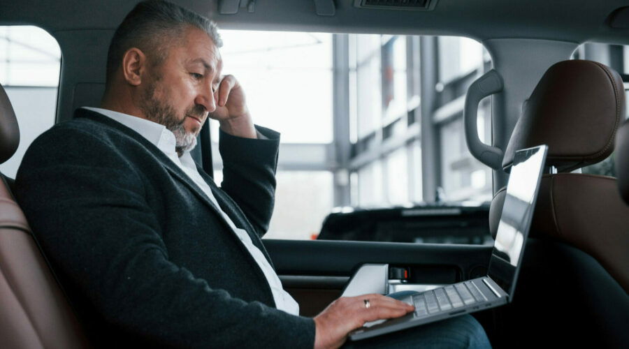 Side view. Working on a back of car using silver colored laptop. Senior businessman.